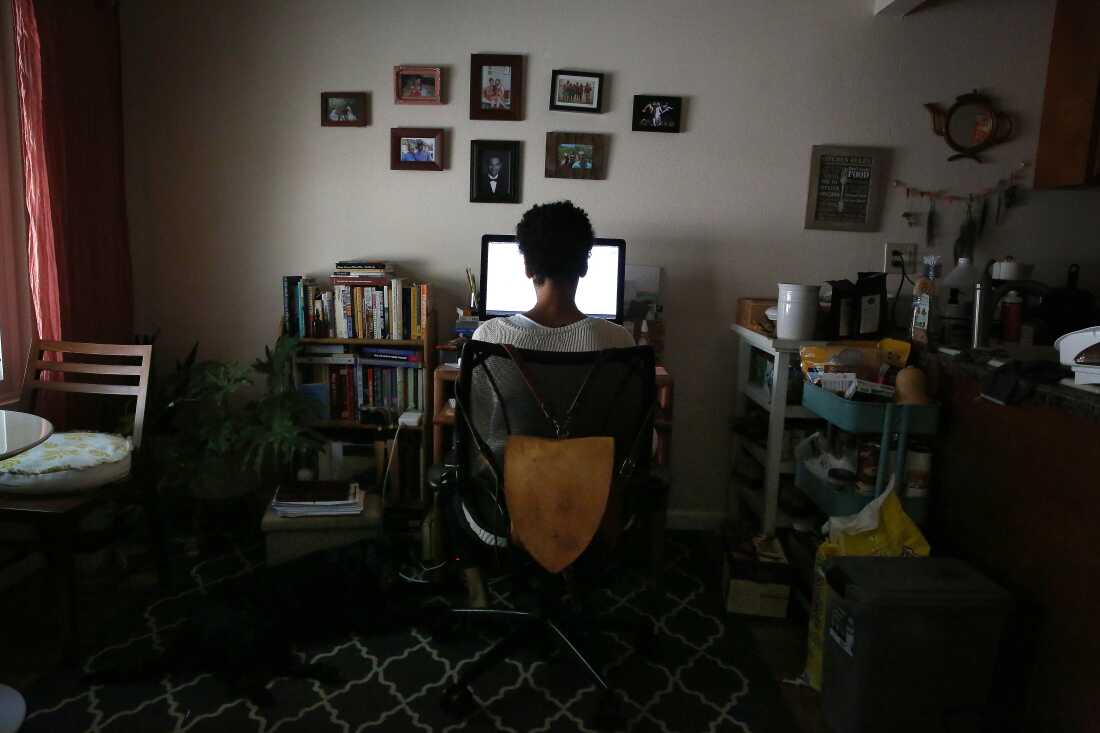 A woman is shown from behind at a sitting at a desk facing a computer screen. An array of small, framed photographs are on the wall above the computer.