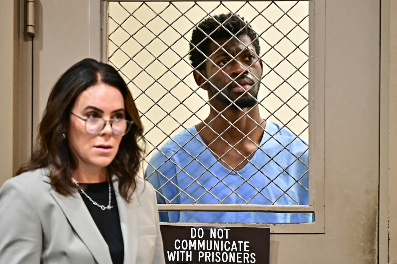 A woman in a suit stands outside of a door with a barred window. On the other side of the window a man in a blue jumpsuit looks on in concern. Below the window is a sign bearing the words 'Do not communicate with prisoners.'