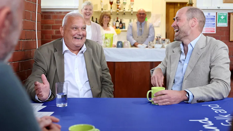 Prince William, to the right, and a man to the left sit at a table with a blue tablecloth. The man to the left has very short grey hair and is smiling, wearing a khaki suit jacket and white shirt. Prince William is turned to look at him and is laughing. He has his left hand around a green mug placed on the table and is wearing a beige jacket and pale blue shirt.  