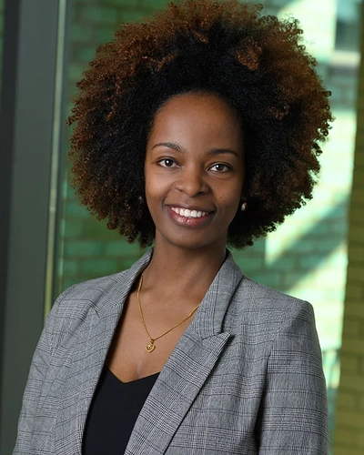 Maji Hailemariam Debena, a mental health epidemiologist and implementation scientist in Michigan State University’s Charles Stewart Mott Department of Public Health, smiles while standing indoors in front of large windows, wearing a gray plaid blazer and black top.