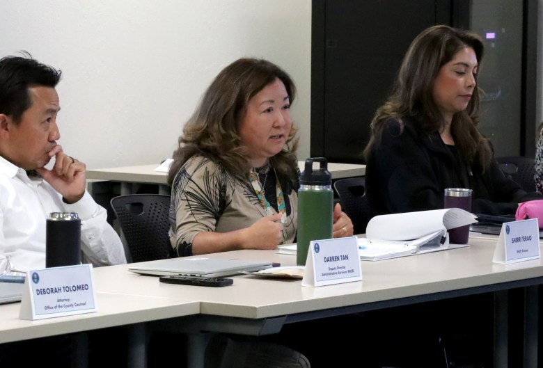 Four individuals are seated at a conference table with nameplates, engaged in a meeting or presentation.