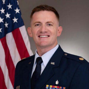 A man in a dark blue uniform and tie with pins on his lapels that read, “U.S.,” and colored stripes below a silver medal on his left breast pocket, stands smiling with the United States flag over his right shoulder.