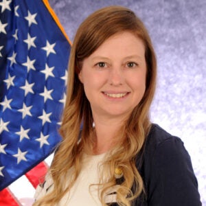 A woman with long, light brown hair wearing a blue blazer and white top is smiling in front of a United States flag.