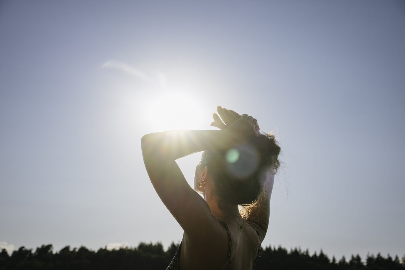 A photo of the back of a woman's head holding her arms above her head and looking into the sunlight.