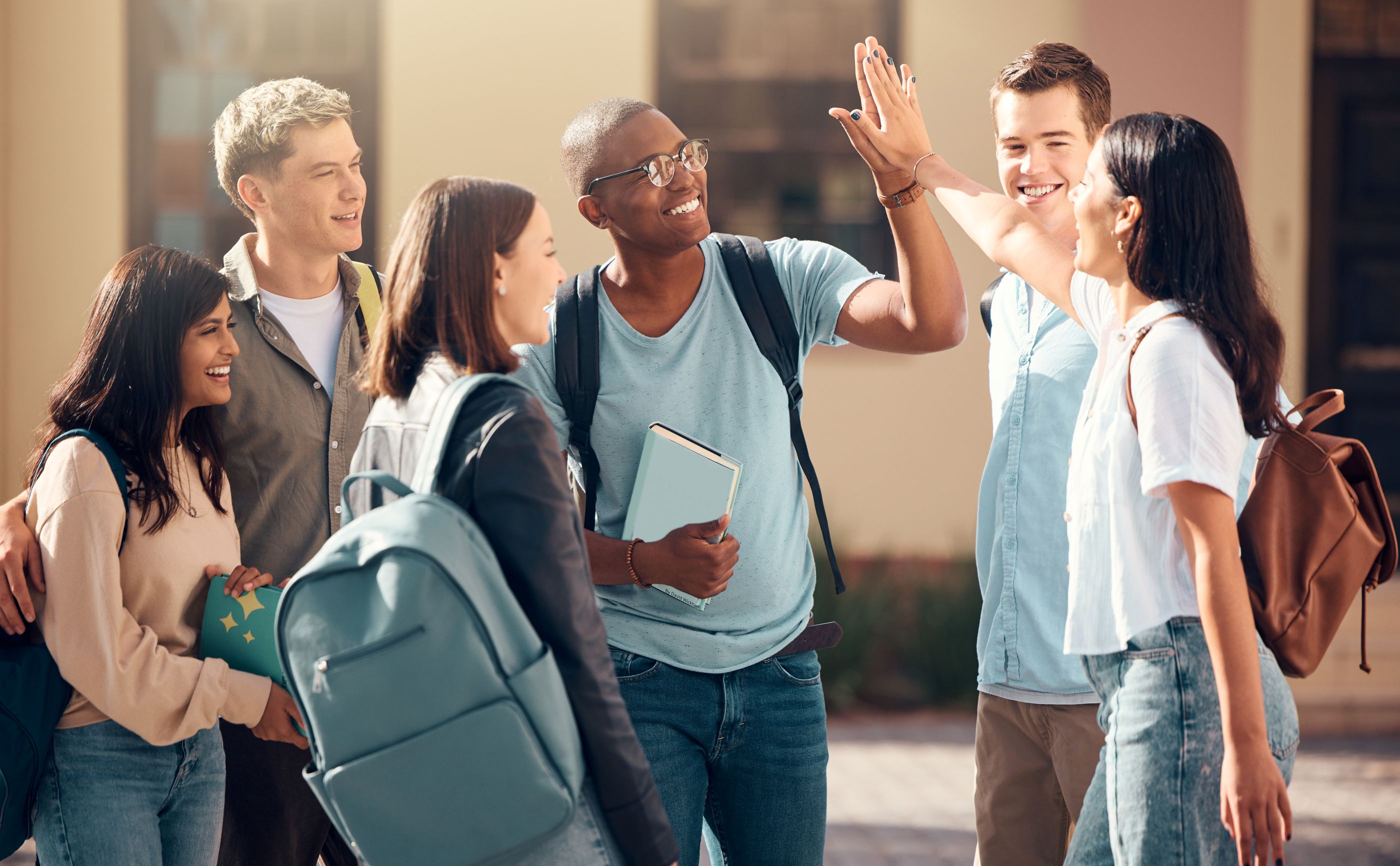 Six college students gather outdoors on campus, smiling and chatting. Two students share a high-five while others look on, creating a welcoming scene of friendship, support, and community