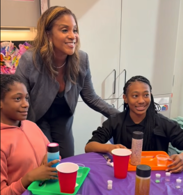 Woman in black dress and gray blazer standing behind two teens seated at table with her hands on their shoulders, all three smiling