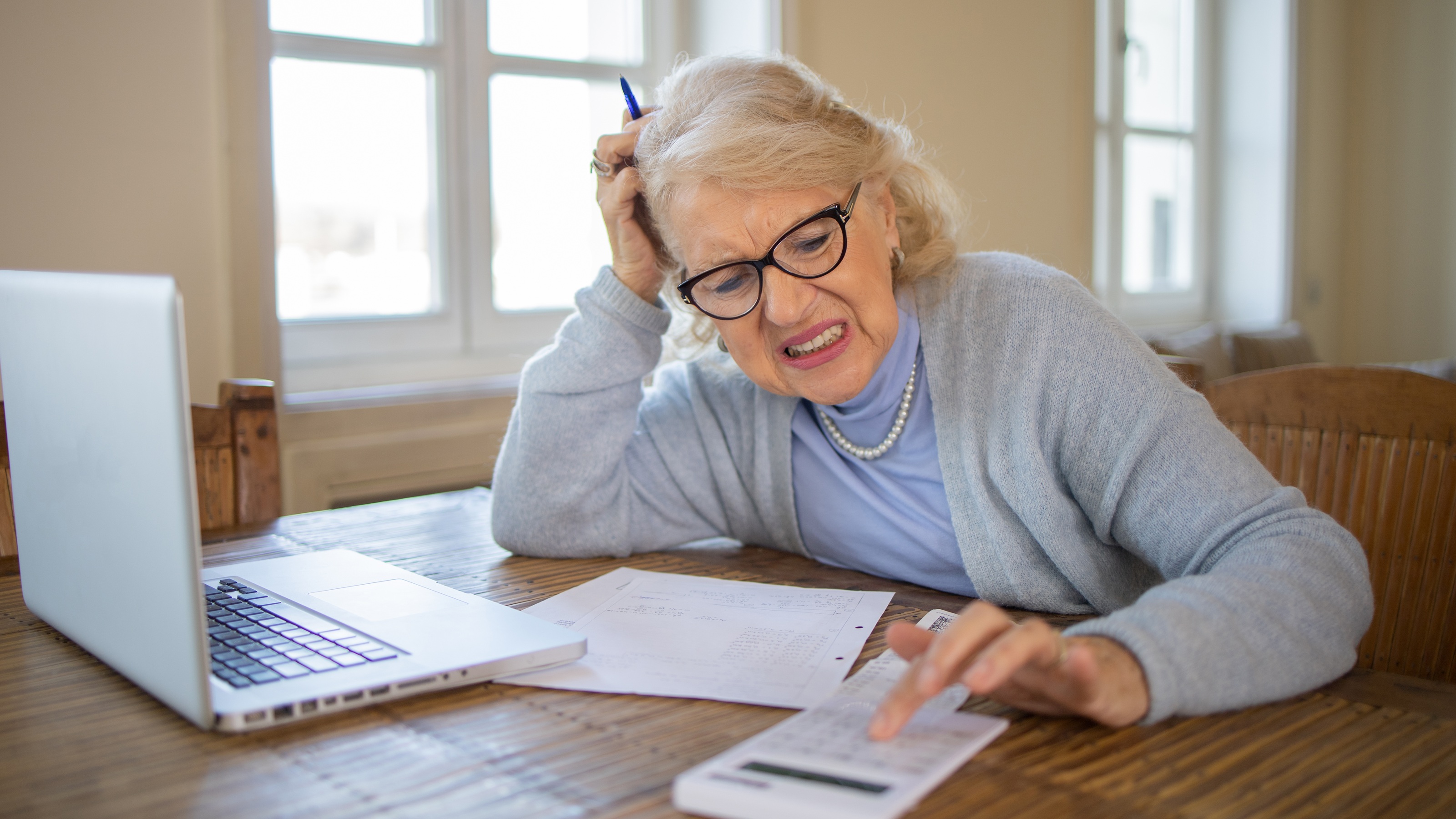 An older woman grimaces as she uses a calculator at her kitchen table.