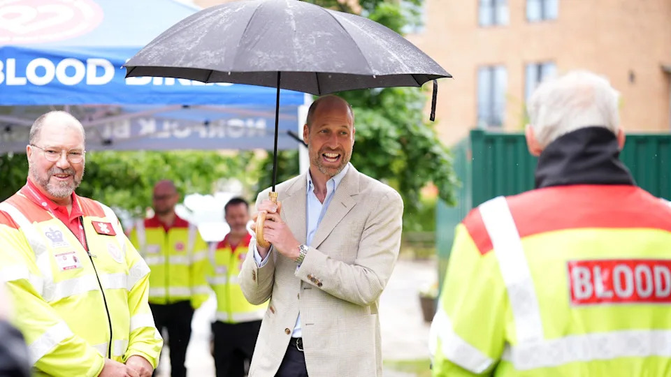 Prince William, dressed in dark trousers, a pale jacket and pale shirt, holds a black umbrella as he talks to men in hi-vis jackets. One has "BLOOD" written across the back of his hi-vis 