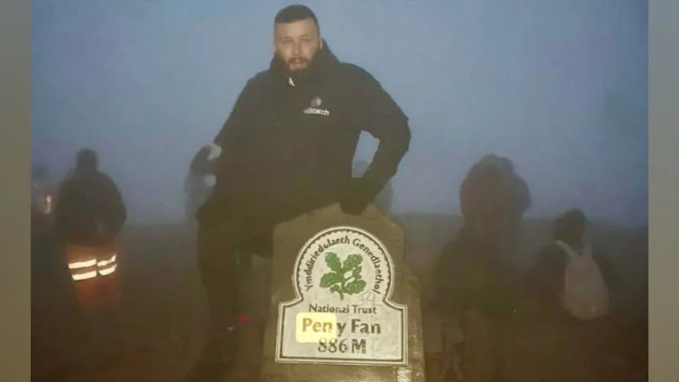 Lee, wearing a black coat, at the summit of Pen y Fan. He has his hand on a stone marker which marks the mountain's height at the top
