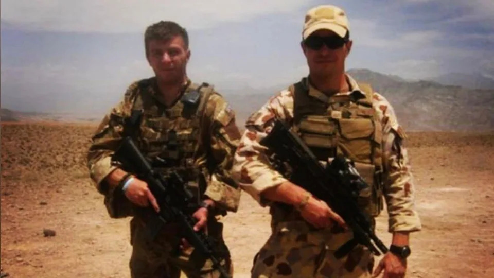Two men in camouflage gear, holding guns, standing in a desert. One has a cap and sunglasses on