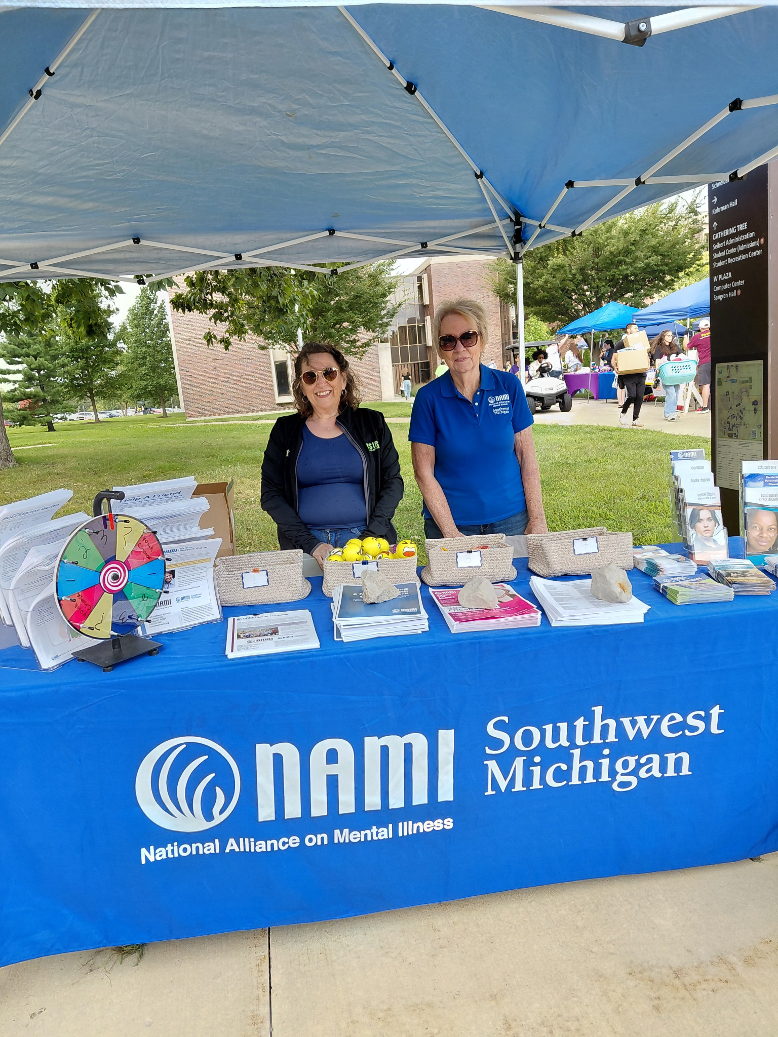 Two women stand at a booth in a park behind a table covered in pamphlets. A blue tablecloth bears the words "National Alliance on Mental Health Southwest Michigan."