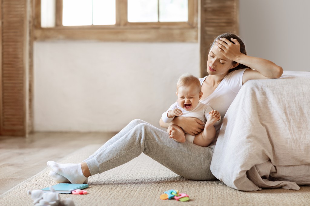 A stressed mother sits on the floor with her hand on her head, holding a crying infant.